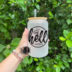 May include: Frosted glass tumbler with a bamboo lid and straw. The tumbler features the phrase "The Hell I Won't" in black script within a circular design, with a cowboy hat graphic. The background is a lush green foliage.