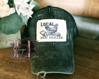 May include: Distressed black baseball cap with a mesh back. The front patch reads "LOCAL EGG DEALER" with a graphic of a hen and eggs. The cap has a pre-worn look with distressed details.