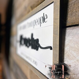 May include: A rectangular wooden sign with a white background and black lettering. The sign reads "Love the people". The sign is framed in a light brown wood and is mounted on a wooden wall.