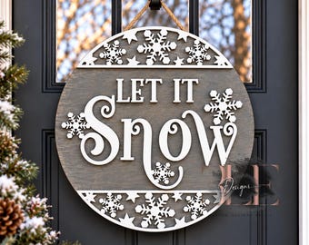 May include: A circular wooden door sign with a dark gray finish featuring the white text LET IT SNOW in a mix of block and script fonts. The sign is decorated with white cutout snowflakes and stars along the top and bottom edges.