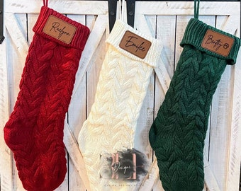 May include: Three cable-knit Christmas stockings in red, cream, and green hang from a wooden beam. Each stocking has a personalized leather patch with a name. The stockings are displayed against a white wooden background.