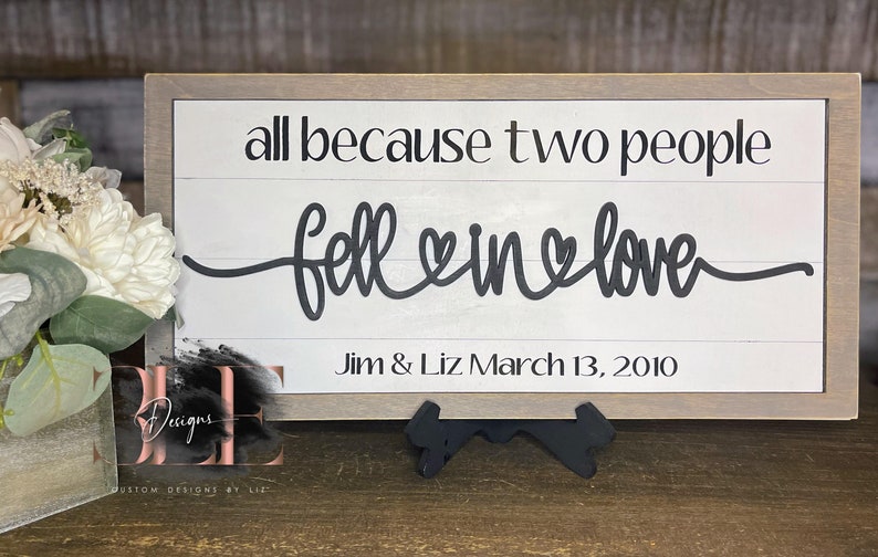 May include: A framed wooden sign with the phrase "all because two people fell in love" in black script. The sign includes the names "Jim & Liz" and the date "March 13, 2010". The sign is a decorative item.