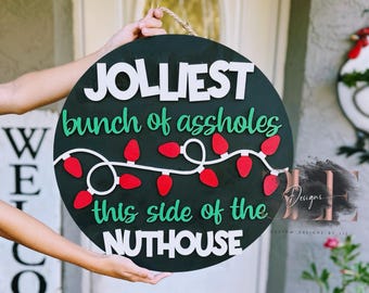 May include: A circular black wooden sign with the text "JOLLIEST bunch of assholes this side of the NUTHOUSE" in white and green. The sign features red Christmas lights. A rope is attached for hanging. The sign is held by a person.