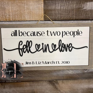 May include: A rectangular wooden sign with a gray frame and a white background. The sign reads "all because two people fell in love" in black script. Below the text is "Jim & Liz March 13, 2010".