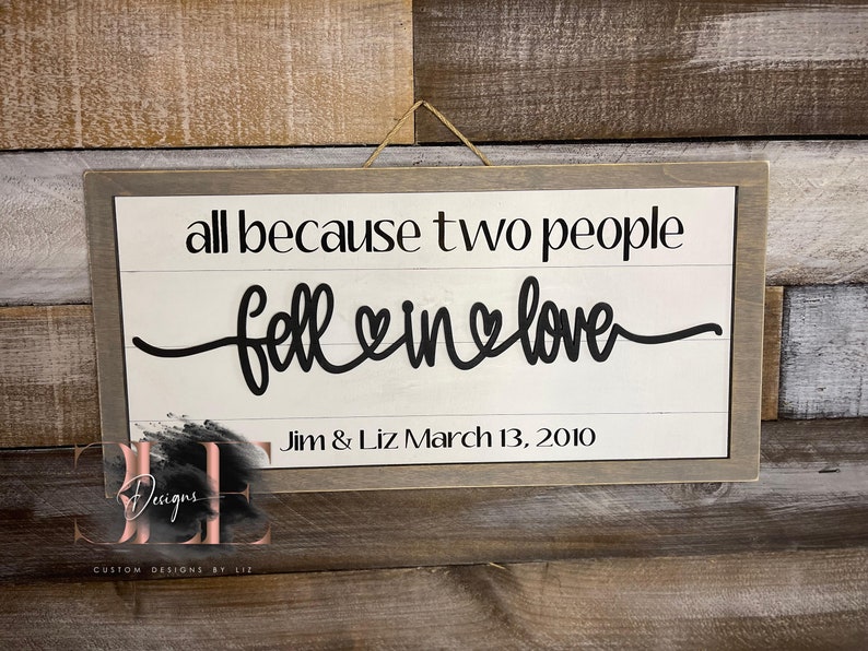 May include: A rectangular wooden sign with a gray frame and a white background. The sign reads "all because two people fell in love" in black script. Below the text is "Jim & Liz March 13, 2010".