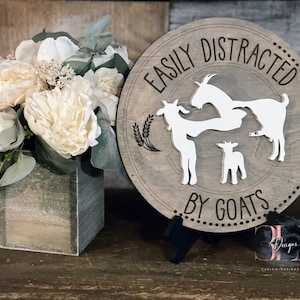 May include: A round wooden sign with the text "EASILY DISTRACTED BY GOATS" and goat silhouettes. The sign is gray with white goats and a black stand. A floral arrangement in a wooden cube is in the background.