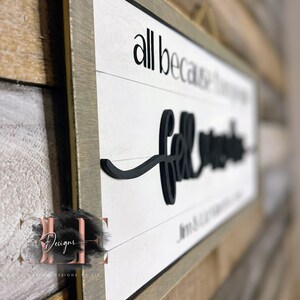 May include: A framed wooden sign with the words "all because two people fell in love" in black lettering on a white background. The sign has a gray frame and is hanging on a wooden wall.