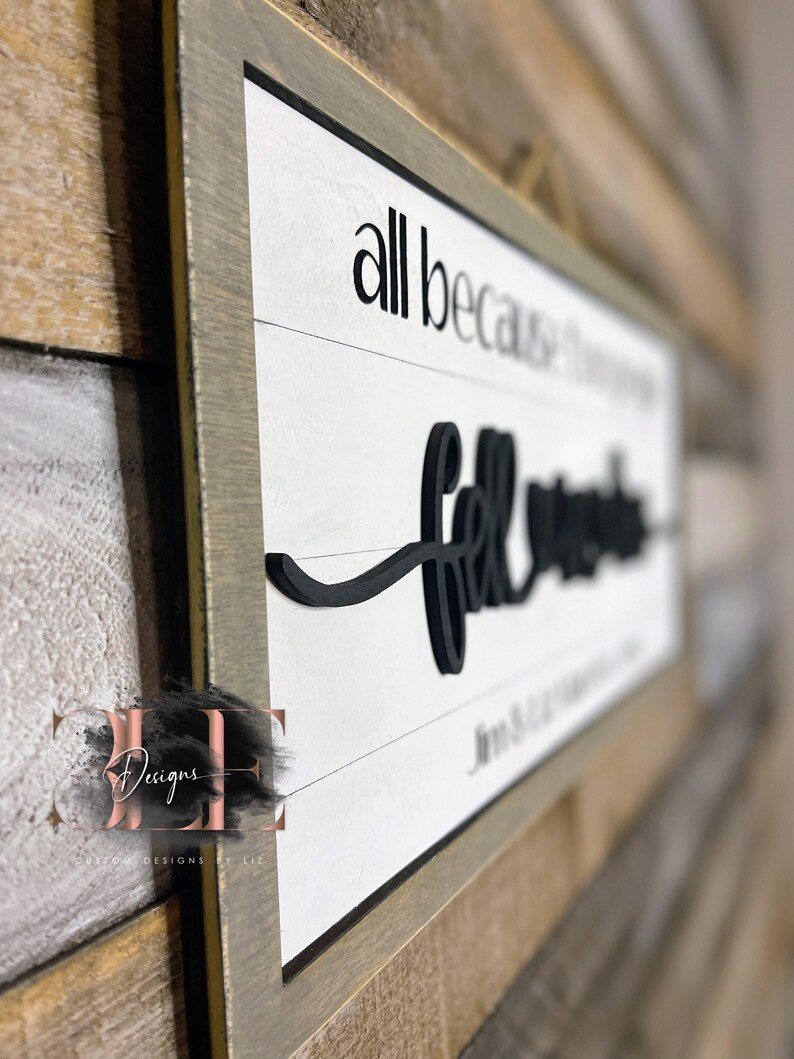 May include: A framed wooden sign with the words "all because two people fell in love" in black lettering on a white background. The sign has a gray frame and is hanging on a wooden wall.