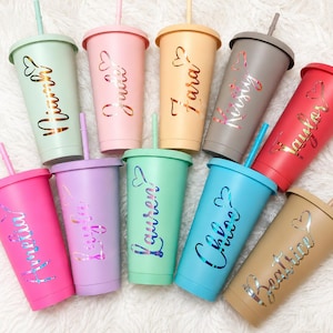 May include: Ten pastel-coloured tumblers with lids and straws, each bearing a name in a cursive font with a heart design. The tumblers are in shades of pink, green, purple, blue, and tan. The image shows a variety of personalised drinkware.