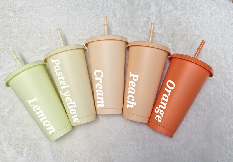 May include: Five reusable tumblers with lids and straws. The tumblers are in various pastel colours, including lemon, pastel yellow, cream, peach, and orange. Each tumbler has a different colour and a white label with the colour name written on it.