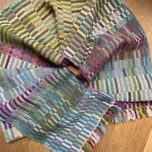 Puede incluir: Una bufanda tejida con un patrón de bloques multicolor en tonos verde, azul, amarillo y magenta. La tela tiene un tejido texturizado e incluye una pequeña etiqueta gris con el texto tinybits&knits. Los extremos tienen flecos.