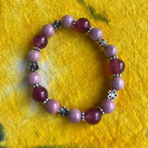 May include: A pink and purple beaded bracelet with silver flower accents. The bracelet is made of round beads and is strung on a clear elastic cord.