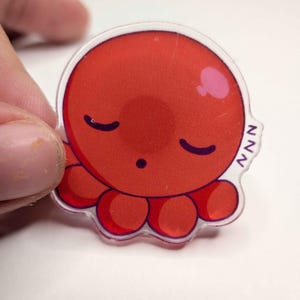 May include: A clear, circular sticker featuring a cartoon octopus with a red body, closed eyes, and a small pink cheek. The octopus has eight red tentacles and the letters "NNN" are printed on the side.