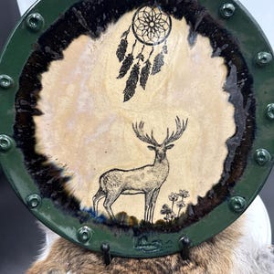 May include: A decorative ceramic plate with a green rim and a deer illustration. The plate features a dreamcatcher design and a deer with large antlers. The plate has a beige center and a dark brown border.