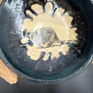 May include: A handmade ceramic bowl with a beaver illustration. The bowl features a dark teal exterior with a light tan interior. The beaver is depicted in a detailed, black and white drawing. The bowl's rim has a unique, textured design.