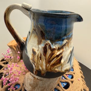 May include: A handcrafted ceramic pitcher with a dark blue glazed interior and a cream-colored handle. The exterior features a leaf design in shades of brown and white. The pitcher is displayed on a wooden base, showcasing its unique, artistic design.
