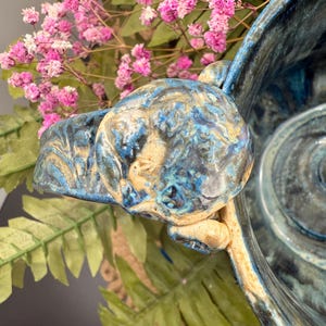 May include: A handcrafted ceramic bowl with a blue and gold glazed finish. The bowl features a unique handle and a textured surface. Pink flowers and green foliage are in the background, adding a natural touch to the composition.