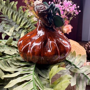 Puede incluir: Un jarrón decorativo de cerámica marrón brillante con un asa y detalles verdes. El jarrón tiene forma bulbosa y se encuentra entre hojas verdes y flores secas. El jarrón es probablemente hecho a mano y añade un toque de encanto rústico.