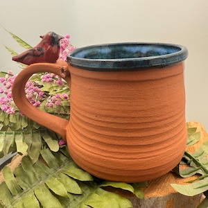 Op de afbeelding: Handgemaakte terracotta mok met een getextureerde buitenkant en een donkerblauwe binnenrand. Een kleine rode vogelbeeldje is bevestigd aan het handvat. De mok wordt gepresenteerd op een houten oppervlak met groen en roze bloemen.