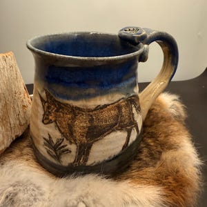 Może przedstawiać: Ręcznie robiony ceramiczny kubek z ilustracją wilka. Kubek ma niebieską obręcz i uchwyt, z beżowym korpusem. Wilk jest przedstawiony w kolorze brązowym i czarnym. Uchwyt ma element dekoracyjny. Kubek stoi na futrzanej powierzchni.
