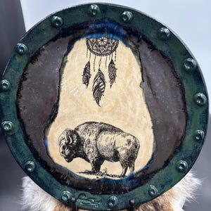 May include: Circular ceramic plate with a dark green and blue rim. The central design features a bison and dreamcatcher in black and white. The plate has a rustic, handcrafted appearance, with visible texture and a natural colour palette.