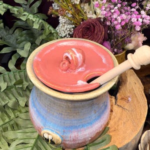 May include: A handmade ceramic butter dish with a red lid and a wooden butter knife. The dish is cylindrical with blue, red, and tan hues. The lid has a knob and a slot for the knife. Decorative flowers are in the background.