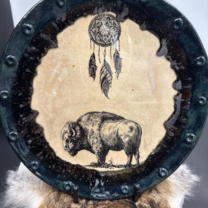 Op de afbeelding: Een ronde keramische plaat met een crèmekleurig midden. Het heeft een zwart-wit illustratie van een dromenvanger en een bizon. De plaat heeft een donkergroene rand met decoratieve accenten.