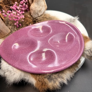 Peut inclure: Une assiette ovale en céramique de couleur mauve mouchetée avec trois sections en creux. L'assiette repose sur une assiette blanche, de la fourrure et un morceau de bois avec des fleurs roses.