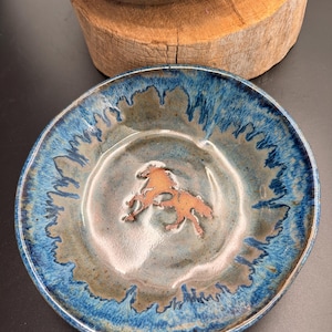 Puede incluir: Un cuenco de cerámica redondo con un esmalte brillante que presenta un borde azul profundo que se transforma en un centro azul más claro y apagado. En el centro del cuenco, hay un relieve de color cobre que muestra dos caballos.