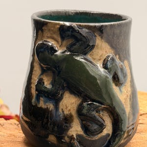 May include: A ceramic mug featuring a raised, dark green lizard sculpture on its exterior. The mug has a glossy, multi-toned glaze in shades of tan, brown, and black, with a vibrant teal interior. The surface texture is uneven and hand-crafted.