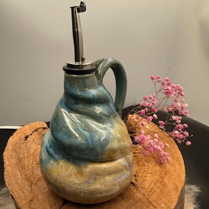 May include: A handmade ceramic oil dispenser with a stainless steel spout. The dispenser has a blue, yellow, and tan swirl design and a curved handle. It is displayed on a wooden slice with pink flowers.