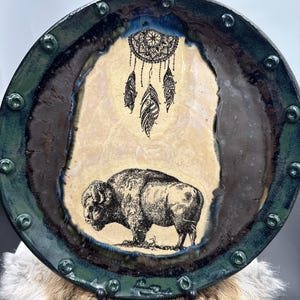 Op de afbeelding: Een ronde, decoratieve plaat met een donkergroene rand en een centraal ontwerp. Het ontwerp toont een zwart-wit illustratie van een bizon en een dromenvanger met veren. De plaat heeft een rustieke, handgemaakte uitstraling.