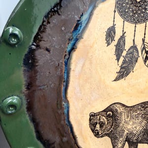 May include: A ceramic plate with a green rim and a central design featuring a bear and a dreamcatcher. The bear is depicted in a detailed, etched style, and the dreamcatcher has intricate patterns. The plate has a rustic, handmade aesthetic.