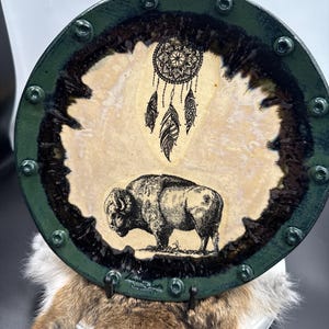 May include: A round ceramic plate with a green rim and a cream-colored center. The center features a black and white illustration of a bison and a dreamcatcher. The rim is decorated with small, silver-colored rivets.