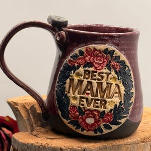 May include: A handmade ceramic mug with a deep plum glaze and a dark base. The front features a circular embossed plaque with the text "BEST MAMA EVER" surrounded by a detailed relief of red flowers and dark green leaves, finished with a curved handle.