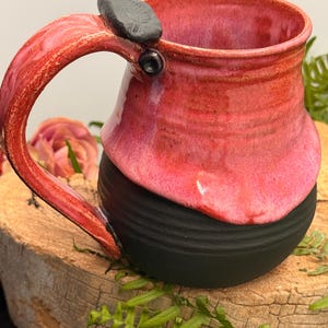 May include: A ceramic mug featuring a two-tone design with a glossy, vibrant pink upper section and a matte black, ribbed lower base. The handle is finished in the same pink glaze and includes a small, dark, textured accent piece at the top.