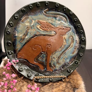 Pode incluir: Prato de cerâmica com um desenho de coelho marrom no centro. O prato tem uma borda verde escura com pontos em relevo e um interior esmaltado azul e marrom. O prato é exibido em um suporte decorativo.