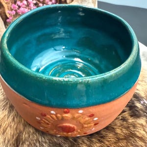 Peut inclure: Un bol en céramique fait à la main avec un intérieur turquoise et un bord vert sarcelle. L'extérieur présente une base en terre cuite avec un motif de soleil en blanc et rouge. Le bol est une pièce de poterie unique.