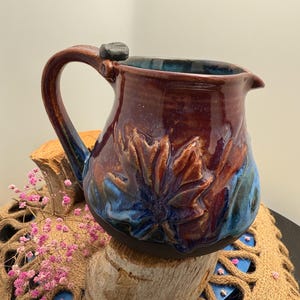 May include: A handmade ceramic pitcher with a floral design. The pitcher features a rich, glossy glaze in shades of burgundy, blue, and brown. The handle is sturdy, and a small, dark stone adorns the top. The pitcher is a decorative and functional piece.