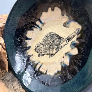 Puede incluir: Un cuenco de cerámica hecho a mano con una ilustración de castor. El cuenco presenta un exterior verde azulado oscuro con un interior color crema y un diseño detallado de castor en blanco y negro. El borde del cuenco tiene un acabado brillante.