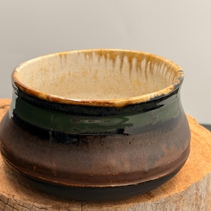 May include: A ceramic bowl with a wide, rounded body and a short, flared rim. The exterior features a layered glaze transition from a dark black base to a deep brown middle and a forest green band near the top. The interior is a speckled cream color.
