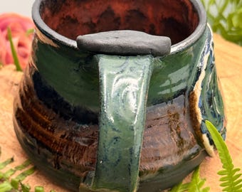 Puede incluir: Una taza de cerámica con un esmalte brillante en tonos verde oscuro, marrón y negro. La taza tiene un asa gruesa y texturizada con un apoyo para el pulgar de color gris oscuro en la parte superior. El interior tiene un esmalte marrón rojizo.
