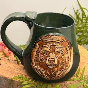 May include: A ceramic mug with a dark green glazed finish and a prominent, raised circular medallion on the front featuring a detailed, brown-toned relief carving of a bear's face. The mug has a thick, curved handle and a smooth, glossy texture.