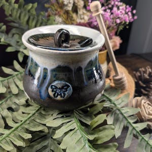 May include: A ceramic honey pot with a lid and a wooden honey dipper. The pot is glazed in black, blue, and white, with a butterfly design. The honey dipper is light brown. The pot is surrounded by green fern leaves.