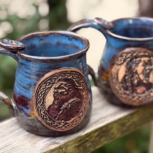 May include: Two blue ceramic mugs with brown accents and copper-colored designs. Each mug features a circular design with a Celtic knot border and a figure inside.