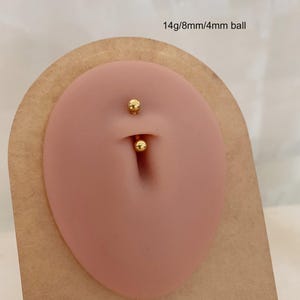 Può includere: Un piercing per ombelico color oro con due sfere alle estremità. La sfera superiore ha un diametro di 8 mm, quella inferiore di 4 mm. Il testo "14g/8mm/4mm ball" è visibile.