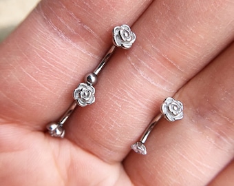 May include: Two silver barbell body jewelry pieces with rose-shaped ends. The jewelry is shown on a hand.