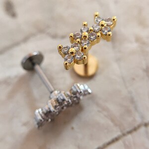 May include: Two gold and silver body jewelry pieces with clear crystal accents. The gold piece features a cluster of three flower-shaped designs with clear crystals. The silver piece features a single flower-shaped design with clear crystals.