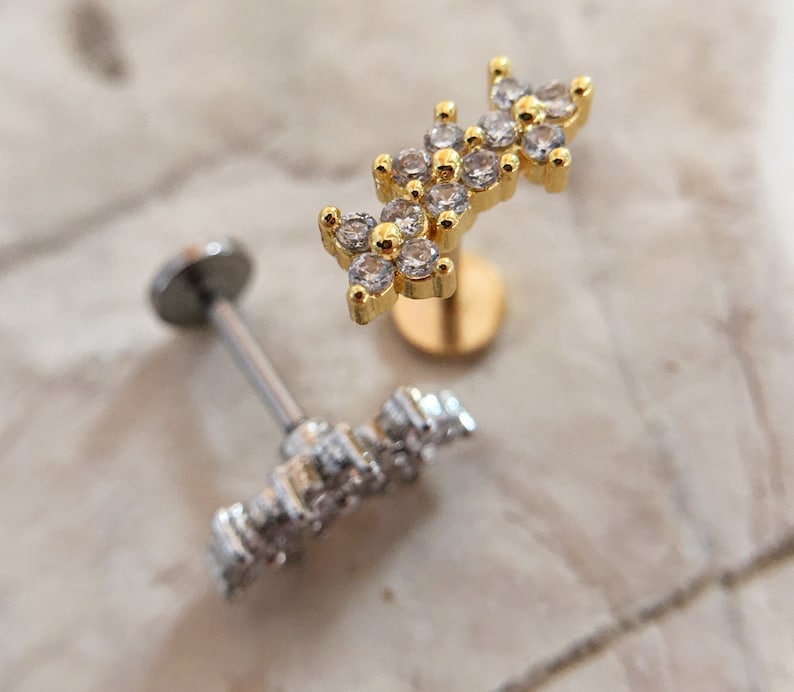 May include: Two gold and silver body jewelry pieces with clear crystal accents. The gold piece features a cluster of three flower-shaped designs with clear crystals. The silver piece features a single flower-shaped design with clear crystals.