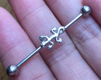 May include: A silver industrial barbell piercing with a lizard charm. The barbell has a straight bar with a small lizard in the center and a ball on each end. The lizard is detailed with visible legs and a tail.
